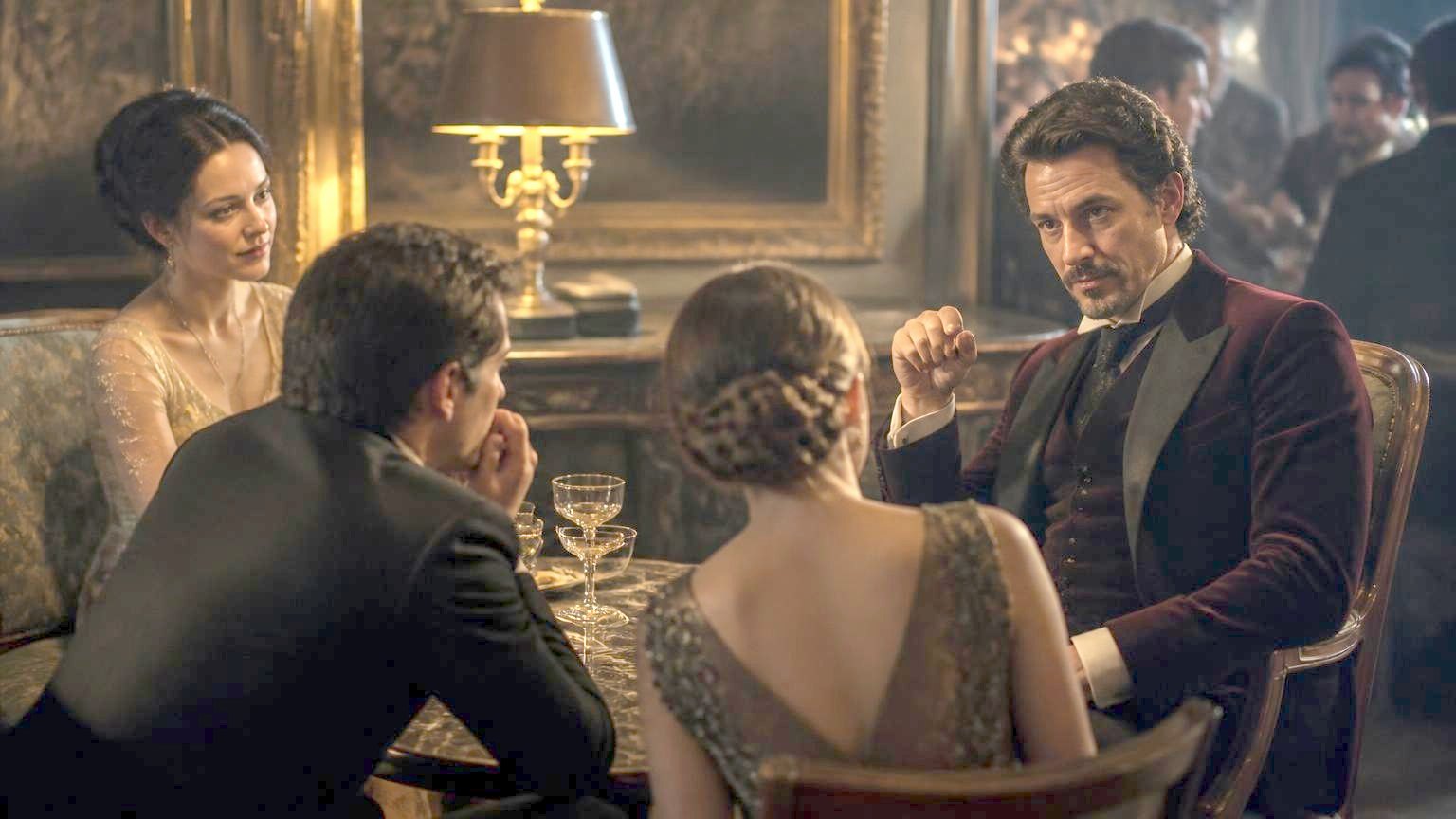 A watchful man in burgundy velvet steers a lamplit conversation with one small deliberate gesture